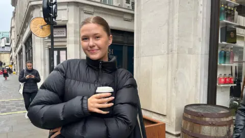 Alice is pictured in a black puffer jacket on the street and holding a coffee cup