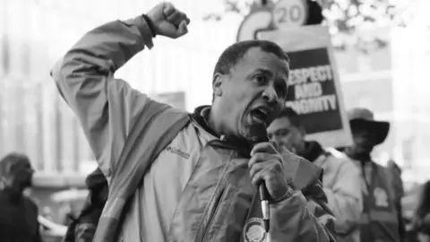 Supplied/IWGB union A black and white picture shows a man, Helio Santos, demonstrating at what appears to be a protest. He has a microphone in one hand and his other hand is raised in a fist. There are signs in the background, and one reads: "Respect and dignity"