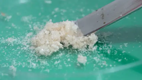 A small mound of white powdery crystals with a spatuala about to take a tiny amount.