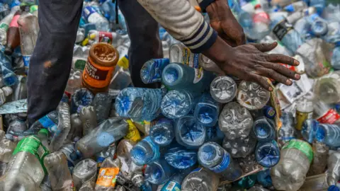 Gerald Anderson/Anadolu/Getty Images Plastic bottles of different colours, clear, blue and orange litter the floor. A black man's hand extends to put some of these bottles into a bag for collection. The lower half of his body is seen, he is wearing blue trousers and a white and blue jacket with striped cuffs.
