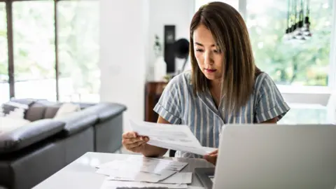 Getty Images A woman looks at her bills while sat in her living room