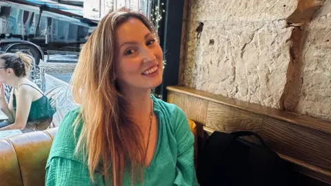 Evie Jay A woman in a green dress sits in front of a window smiling. She is sat in the corner with a stone wall beside her, while a window looks out on to the street behind her.